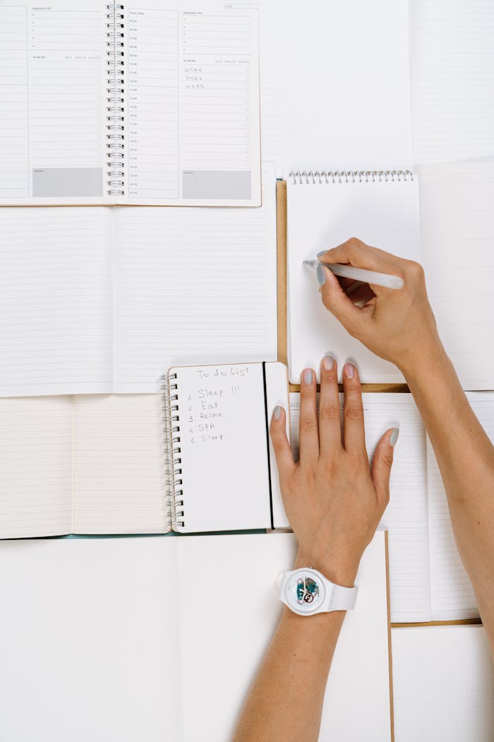 A person writing on a white notepad with a wristwatch, surrounded by open notebooks.