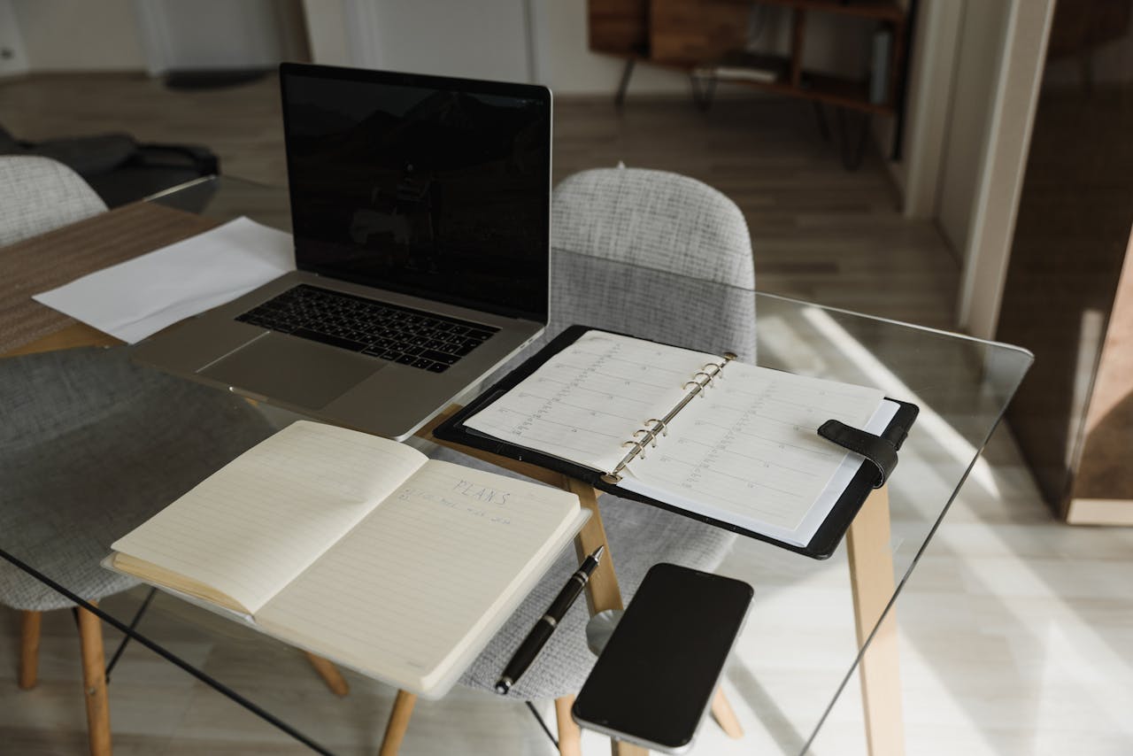 A clean and organized office setup with a laptop, notebook, and planner on a glass desk.